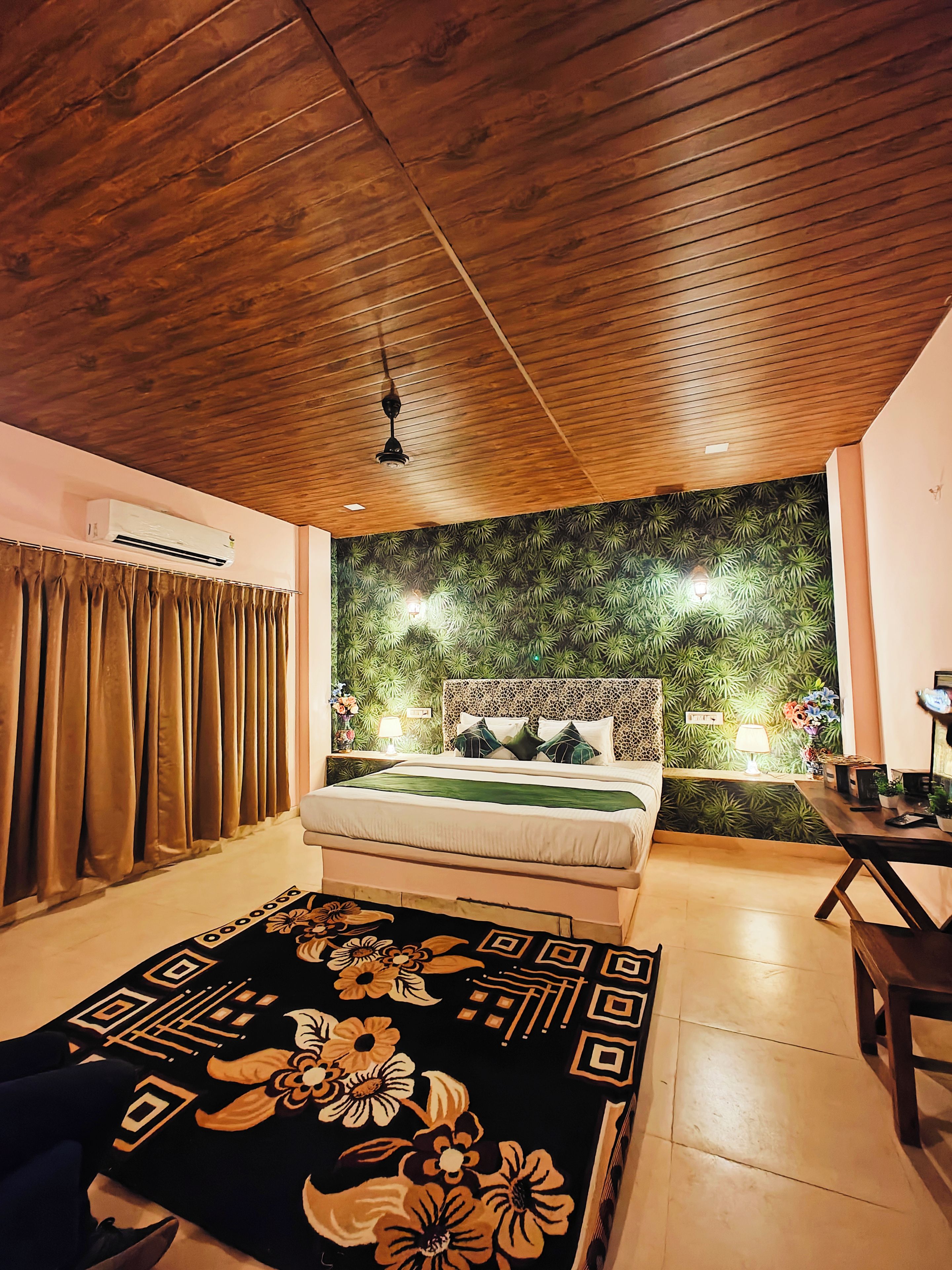 Un dormitorio con techo de madera, cama con cubrecama verde, pared con estampado de hojas y una mesita de noche con lámpara.