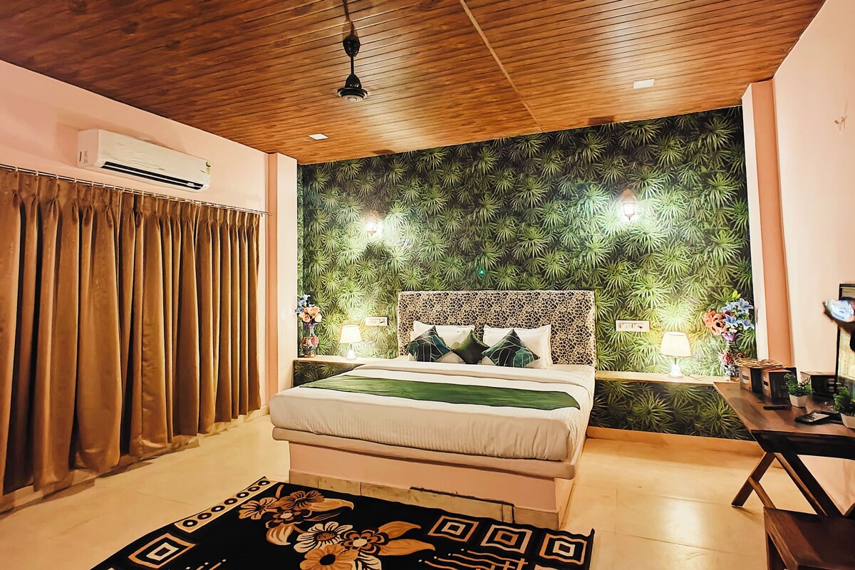 Un dormitorio con techo de madera, cama con cubrecama verde, pared con estampado de hojas y una mesita de noche con lámpara.
