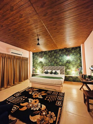 Un dormitorio con techo de madera, cama con cubrecama verde, pared con estampado de hojas y una mesita de noche con lámpara.
