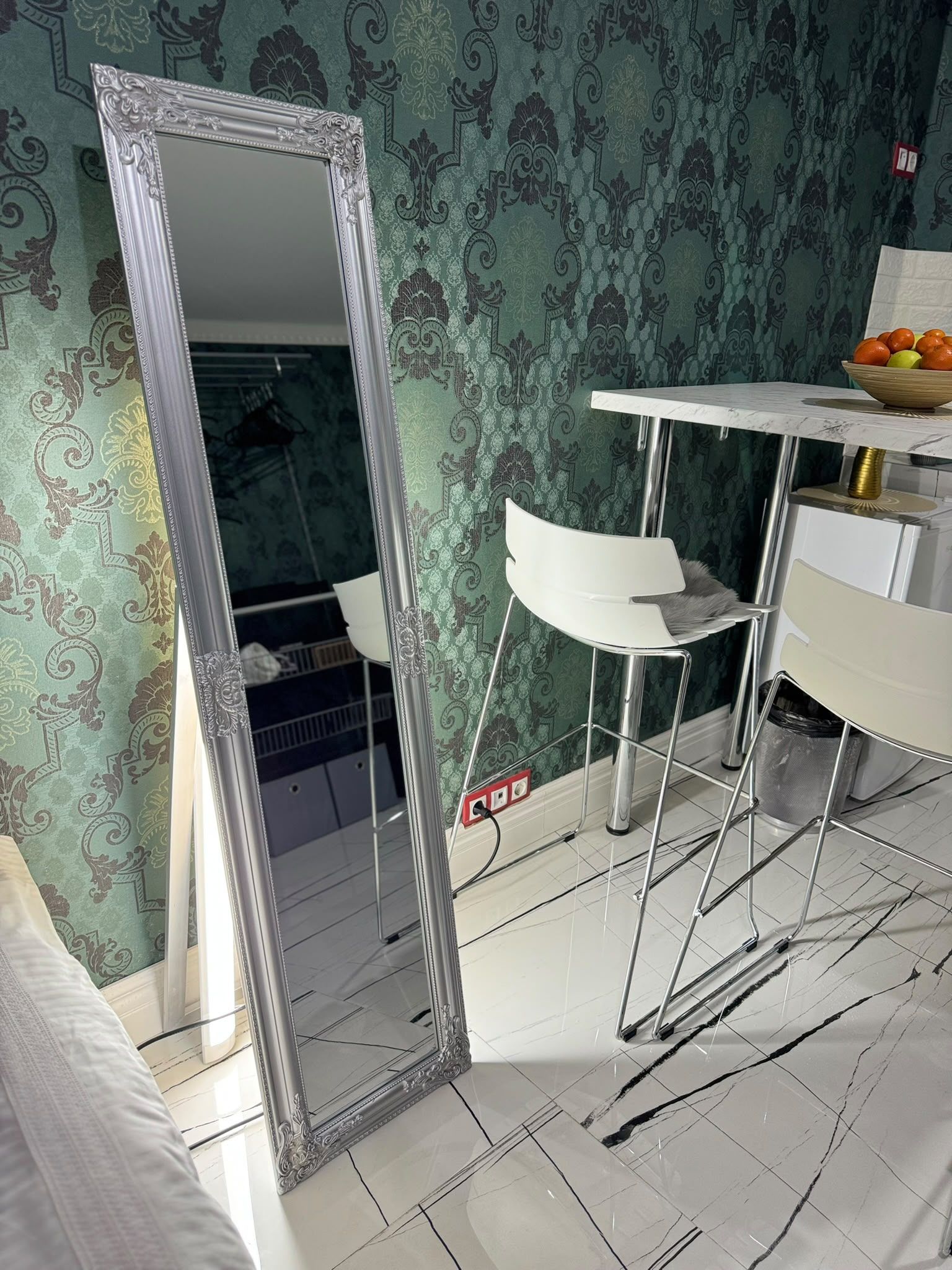 Una habitación con un espejo grande y ornamentado, una silla blanca, una mesita con un cuenco de fruta y papel tapiz estampado.