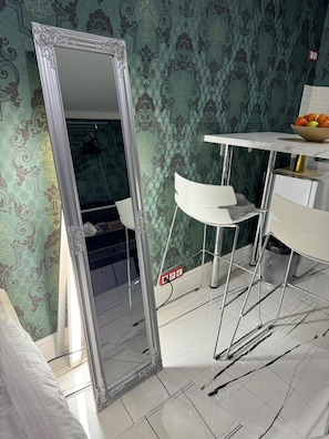 Una habitación con un espejo grande y ornamentado, una silla blanca, una mesita con un cuenco de fruta y papel tapiz estampado.
