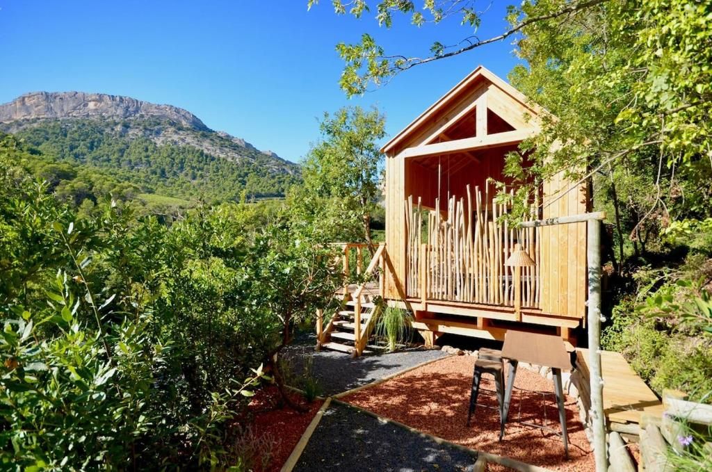 A wooden cabin with a balcony, surrounded by greenery and a mountainous backdrop.