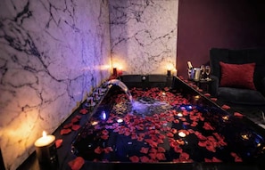 Un área de spa con pared de mármol, fuente con pétalos de rosa, velas y un sofá de color oscuro.