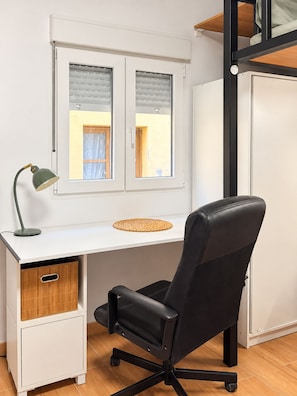 Una habitación con un escritorio blanco, una silla de oficina negra y una ventana con persianas.
