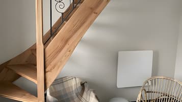 Un escalier en bois avec une rampe en métal noir et un lustre. On aperçoit un fauteuil à carreaux et un fauteuil en osier sur le plancher.