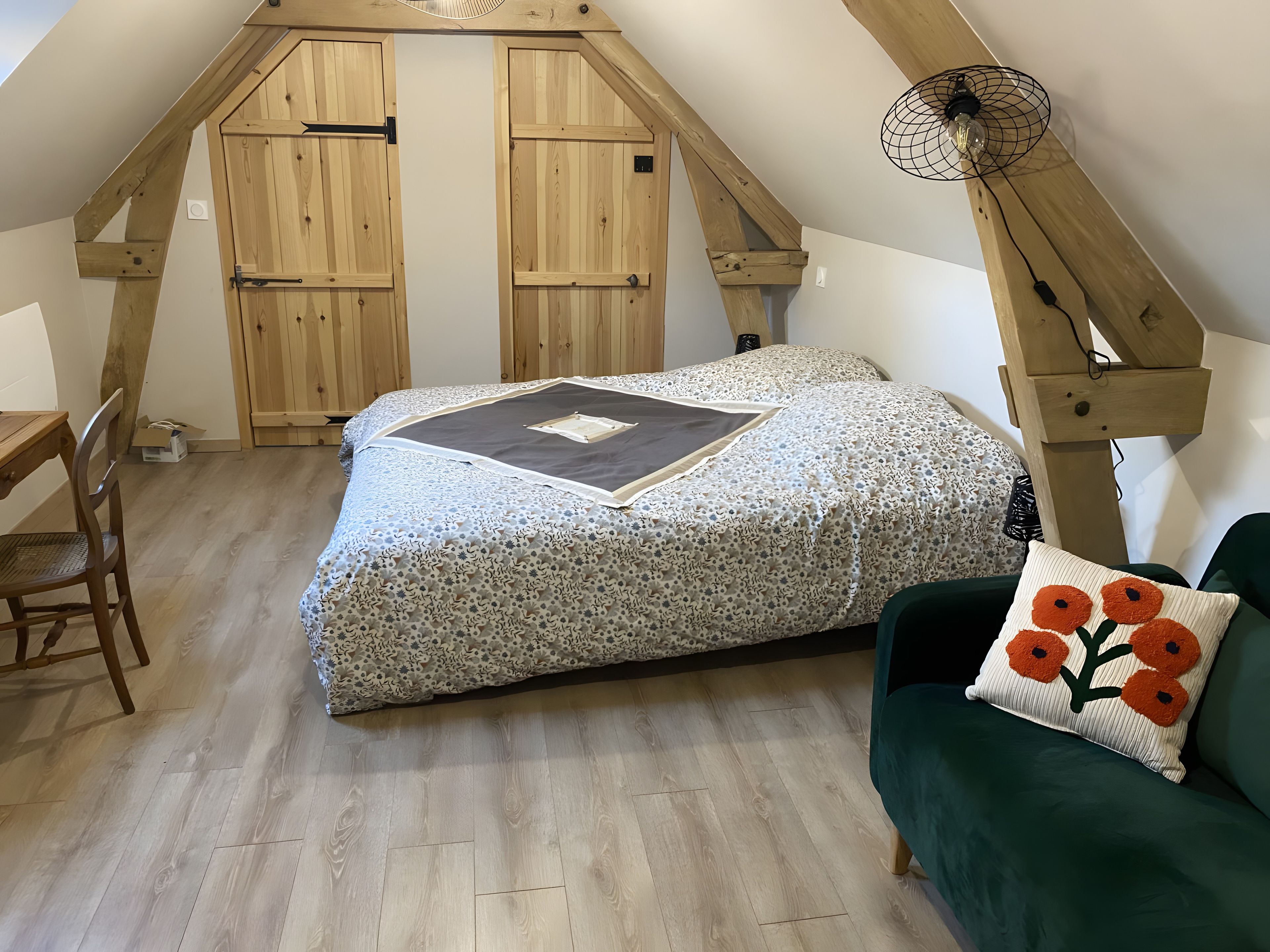 Une chambre avec un plafond en pente, des portes en bois, un lit recouvert d’une couvre-lit à motifs, un canapé vert et une petite table avec une chaise.