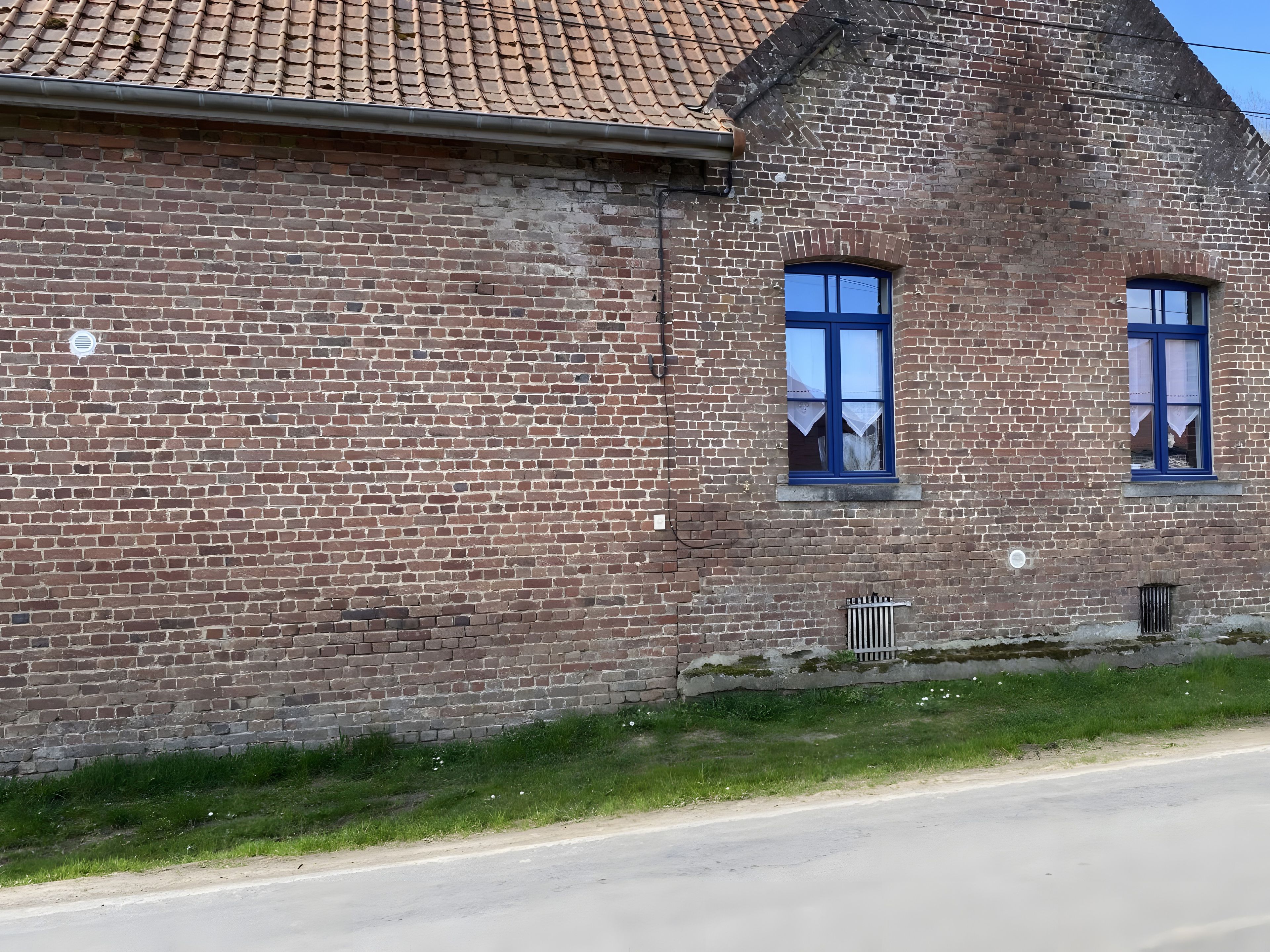 Un bâtiment en briques, doté d’un toit en tuiles et de deux fenêtres aux encadrements bleus.
