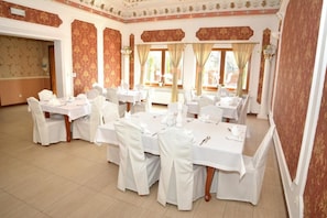 Ein Festsaal mit mehreren runden Tischen, jeder mit weißem Tuch und Stühlen gedeckt, und einer dekorativen Decke mit floralen Mustern.