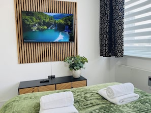 Een slaapkamer met een bed, een aan een houten paneel bevestigde televisie, een plant op een nachtkastje en een raam met gordijnen met een patroon.