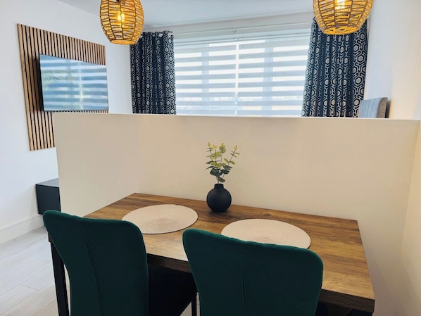 Een eetruimte met een houten tafel, twee groene stoelen, een vaas met bloemen en een televisie aan de muur.