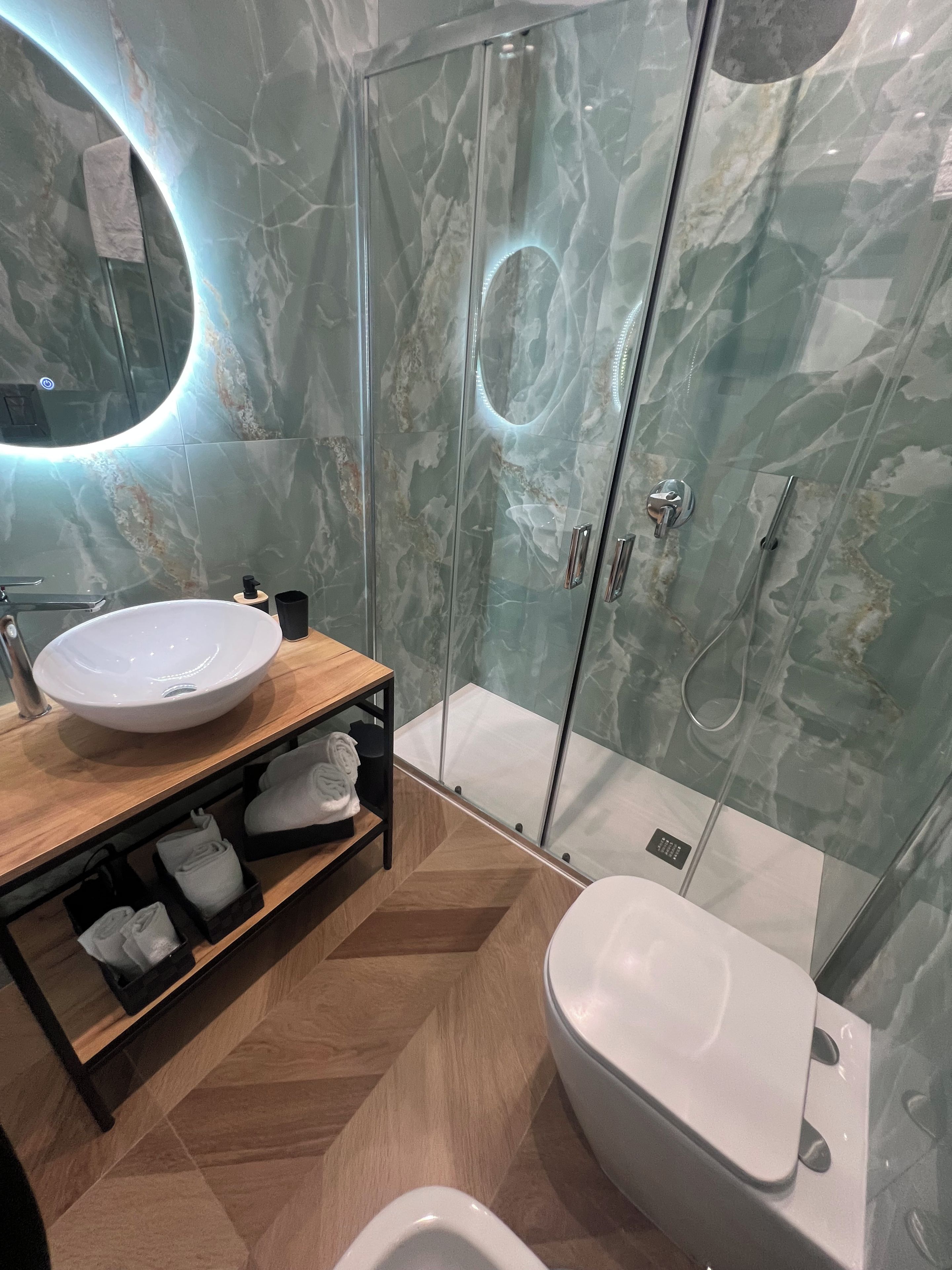 A modern bathroom with a marble shower enclosure, a round mirror, a wooden vanity with a sink, and a toilet.