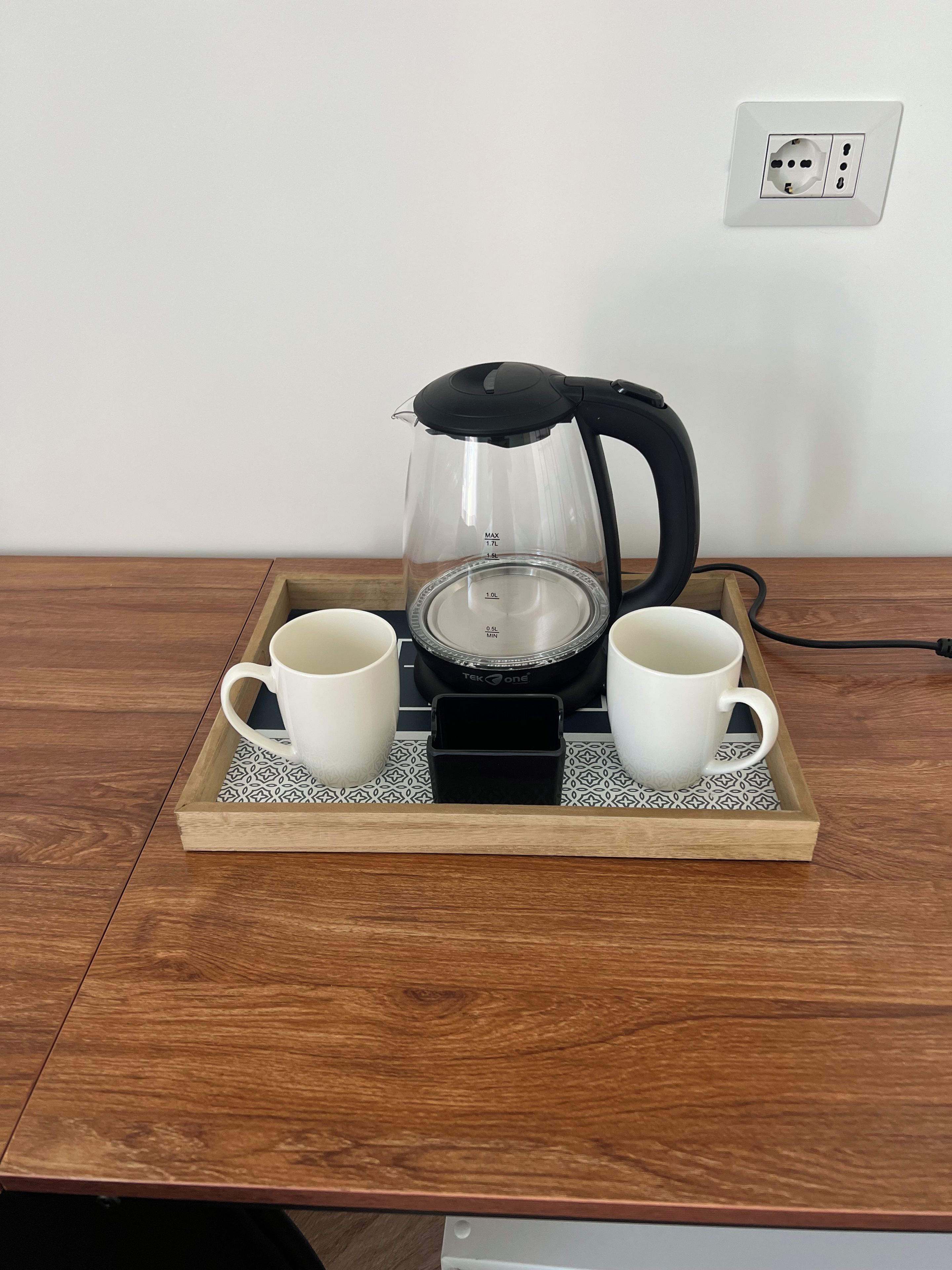 A coffee maker on a tray with two white cups, a power outlet on the wall, and a wooden table.