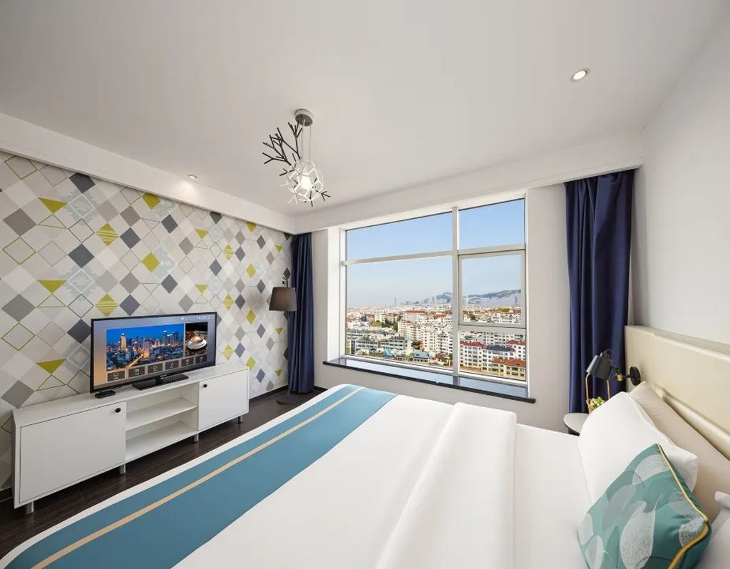 A hotel room with a bed, a TV, a desk, and a large window with a city view.