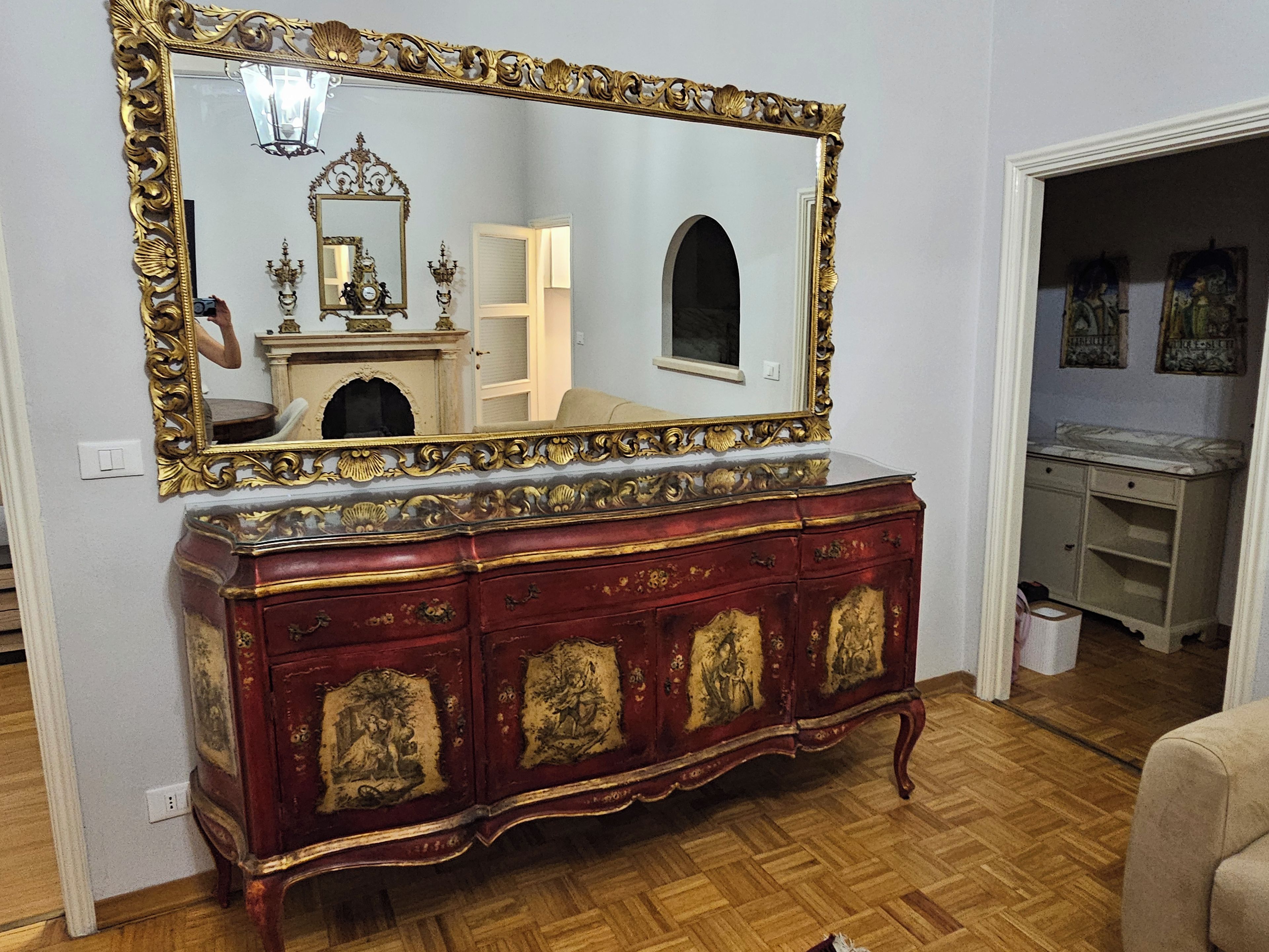 A room with a large ornate mirror, a wooden floor, and a vintage red dresser with gold detailing.