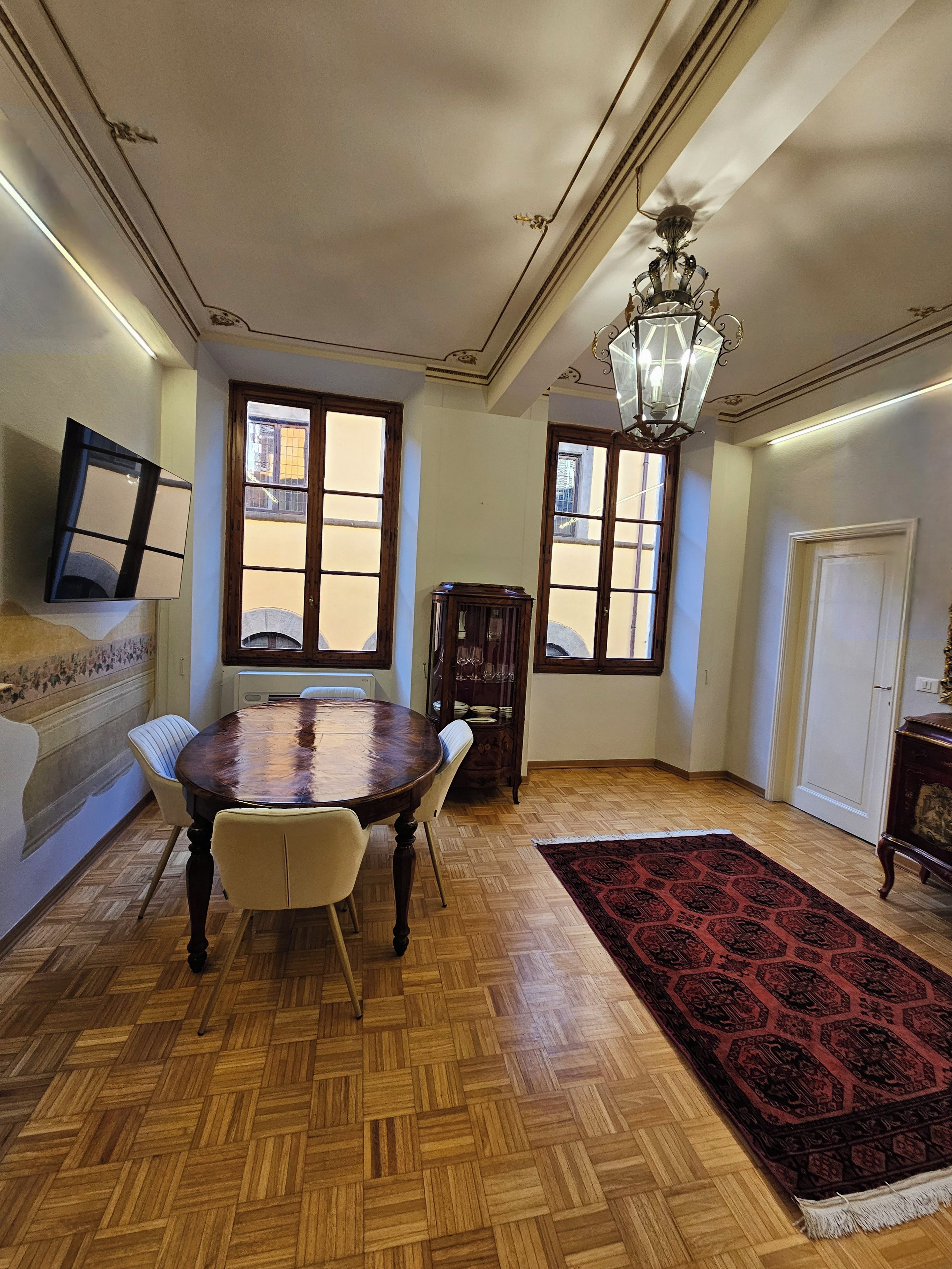 A dining area with a wooden herringbone floor, a round table, and chairs. There is a chandelier, a mirror, and a window with a view.