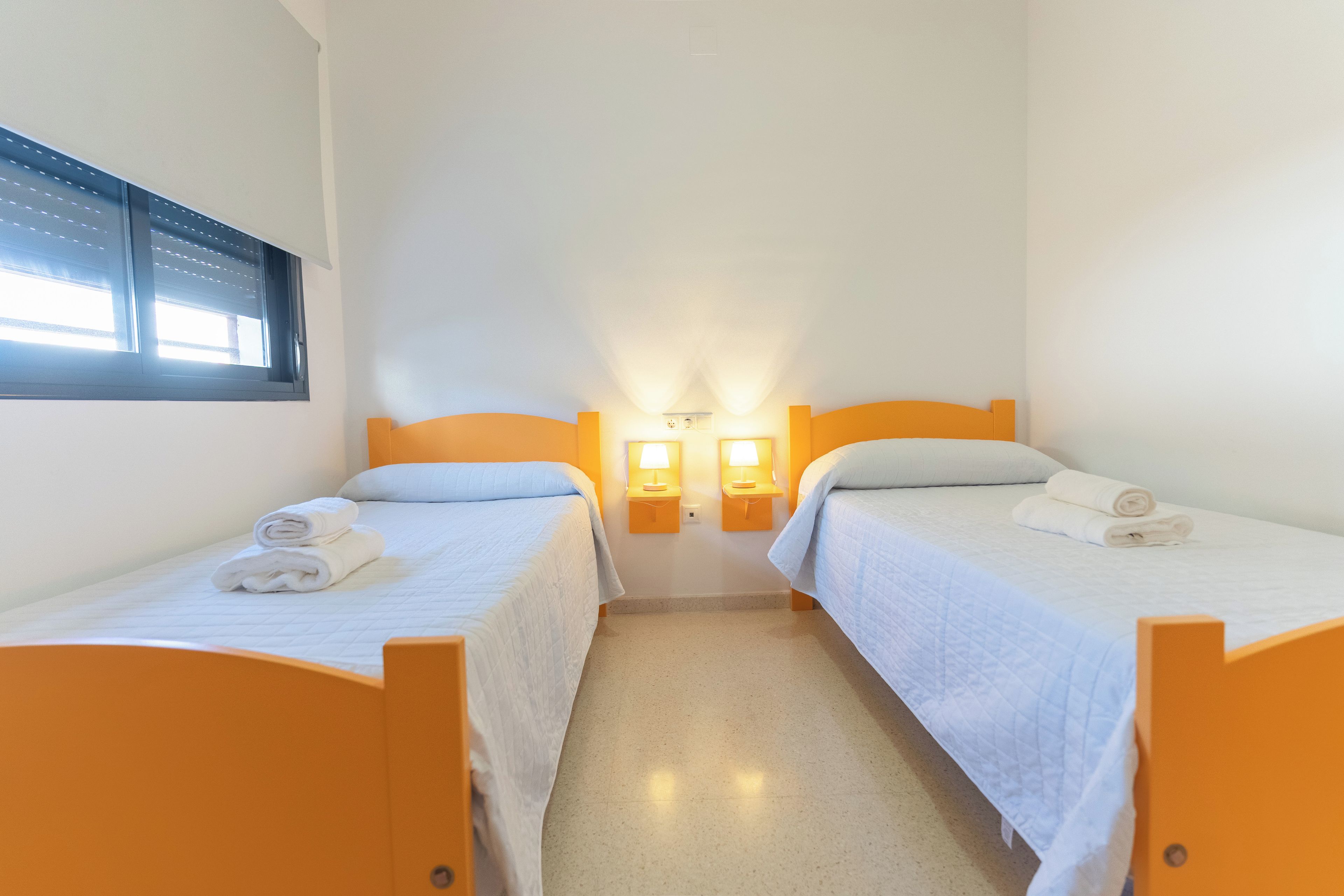 A hotel room with two single beds, each with a white duvet and rolled towels. There are two wall-mounted lamps on either side of the bed.