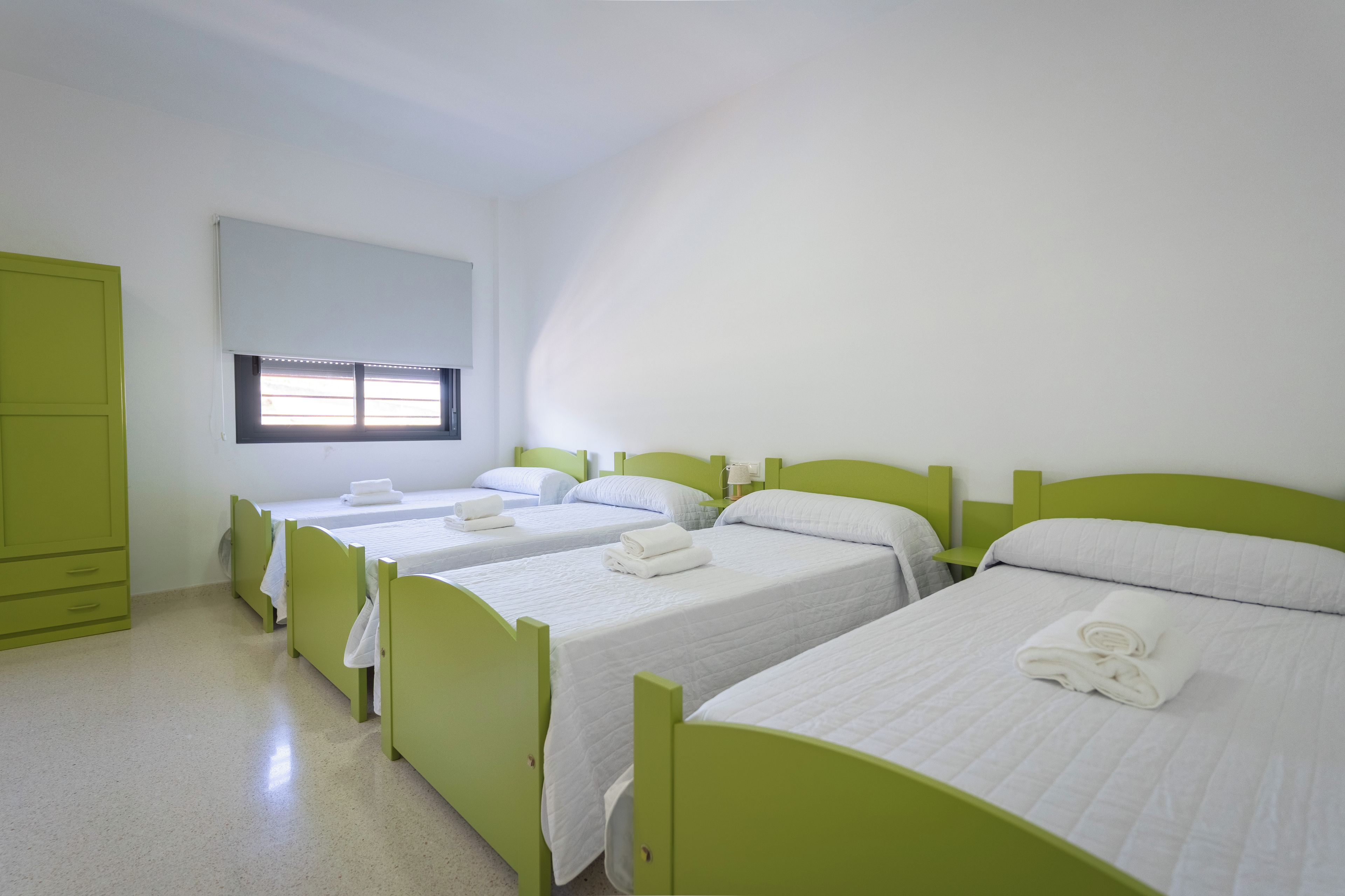 Habitació amb tres llits individuals, cadascun amb capçalera verda i roba de llit blanca. Hi ha una finestra amb persianes, un armari verd i una petita taula de nit amb una làmpada.