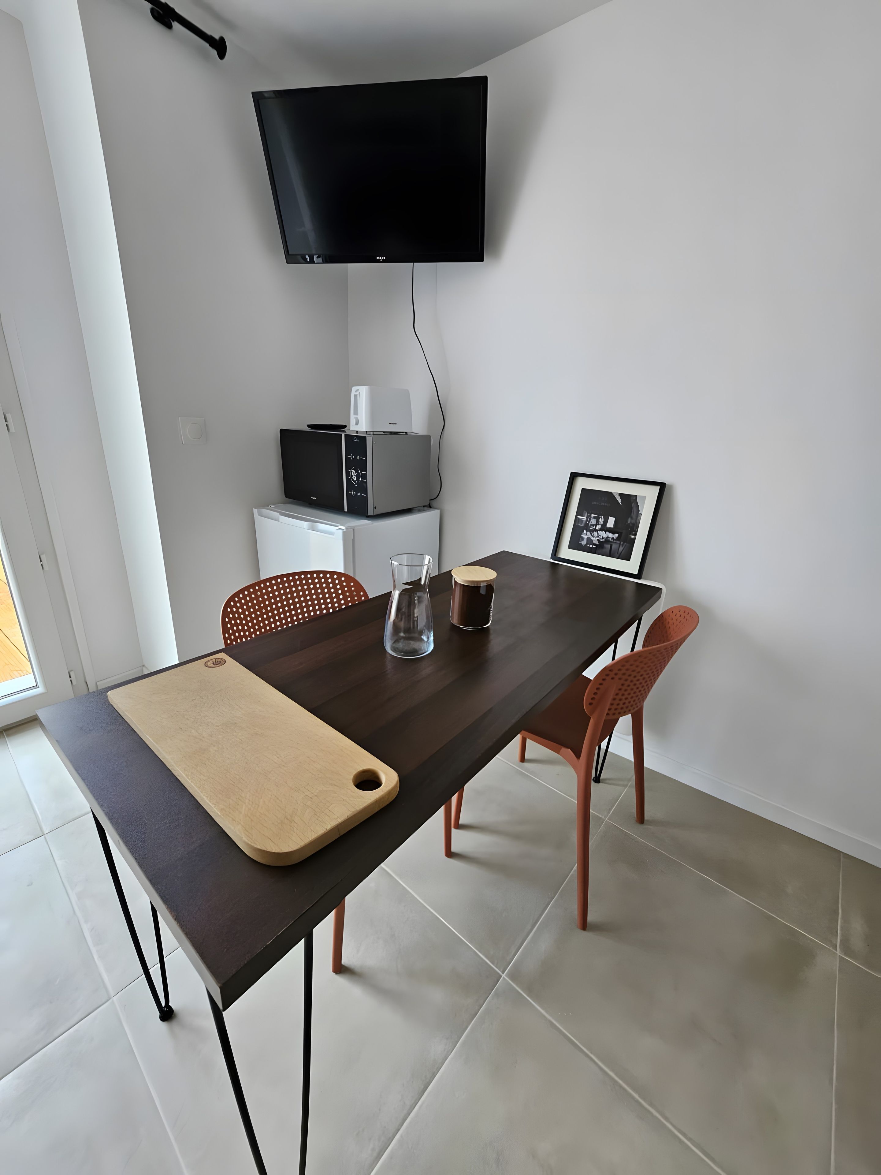 Een eethoek met een donker houten tafel, een snijplank, twee stoelen, een glas en een lijst met een afbeelding.