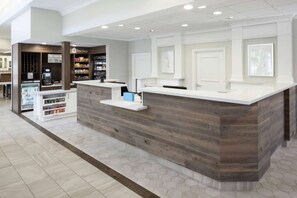 A reception area with a wooden counter, a coffee machine, and a refrigerated display. - Steps to Watters Creek Village + Pool, Bar & Gym (Allen)