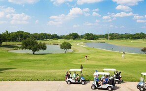 A golf course with multiple golf carts, a pond, and a clear sky. - Steps to Watters Creek Village + Pool, Bar & Gym (Allen)
