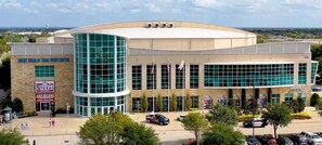 A modern building with a curved glass facade, multiple flags, and a parking lot with several cars. - Steps to Watters Creek Village + Pool, Bar & Gym (Allen)