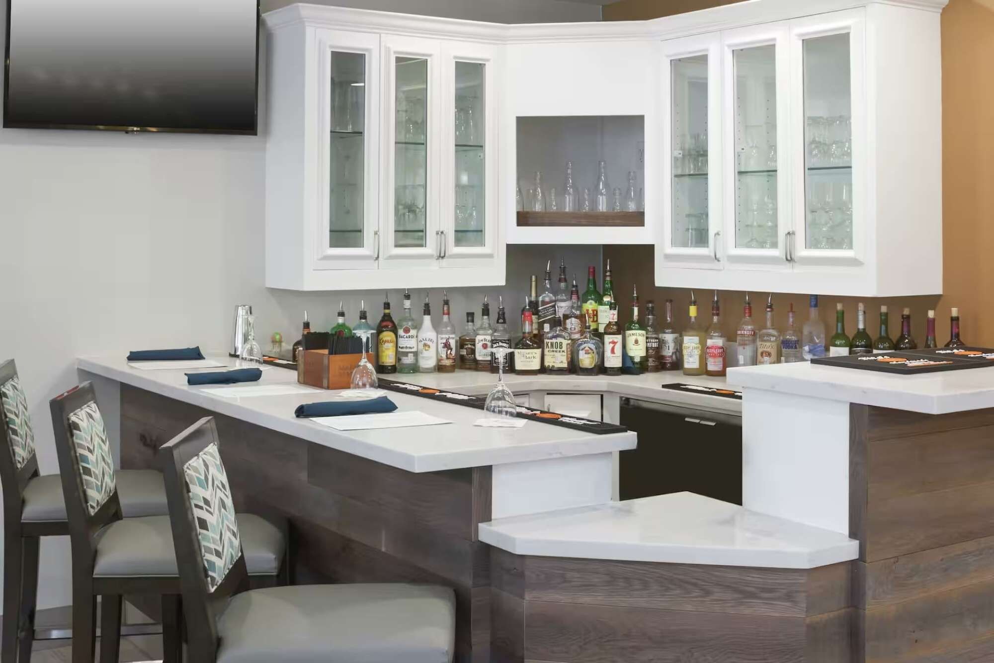 Een moderne keuken met een ingebouwde bar, voorzien van kasten met glazen deuren, een aanrecht met verschillende flessen en een flatscreen televisie die boven de kasten is bevestigd.