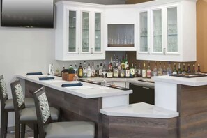 A modern kitchen with a built-in bar area, featuring cabinets with glass doors, a countertop with various bottles, and a flat-screen TV mounted above the cabinets. - Steps to Watters Creek Village + Pool, Bar & Gym (Allen)