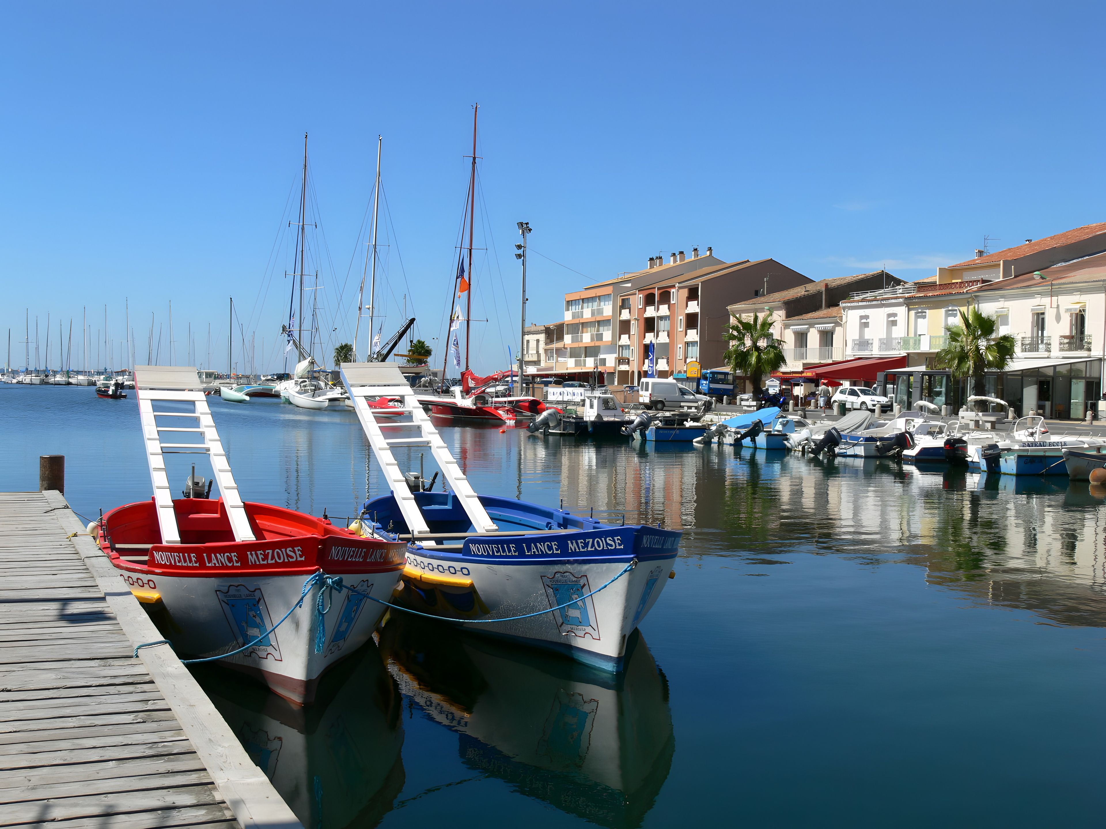 A marina with boats docked, a wooden pier, and buildings in the background.