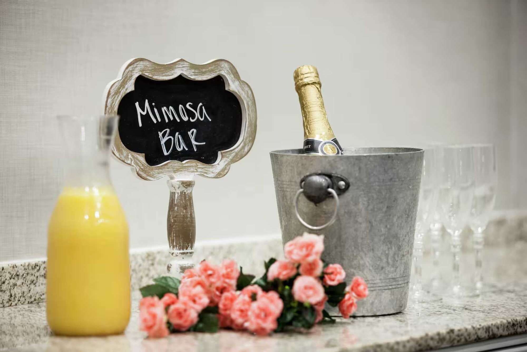 Een barruimte met een bord waarop 'Mimasa Bar' staat, een champagnefles in een metalen emmer en een glas jus d'orange.