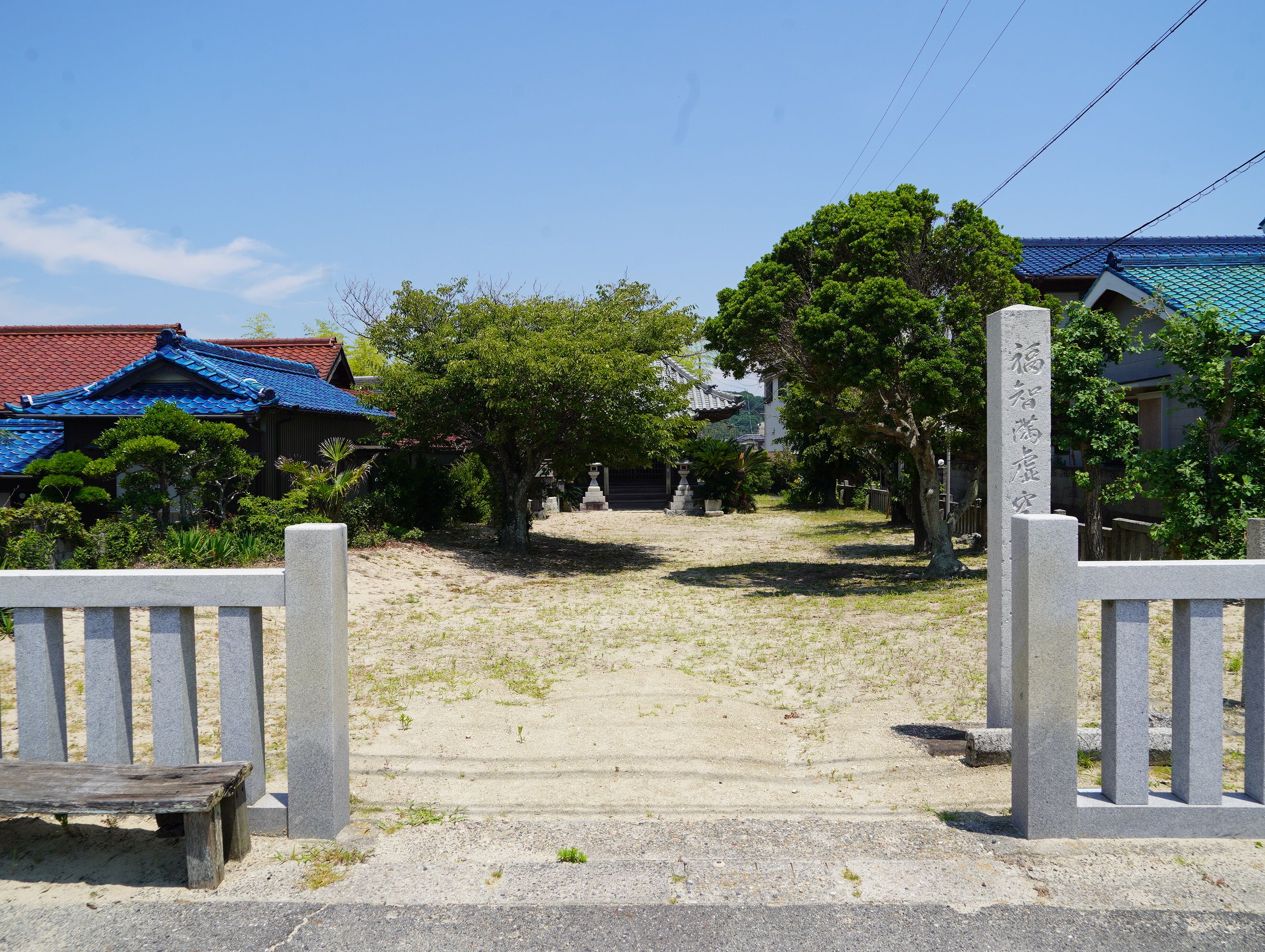 A traditional Japanese gate leading to a courtyard with a stone bench, a stone path, and buildings with blue roofs.