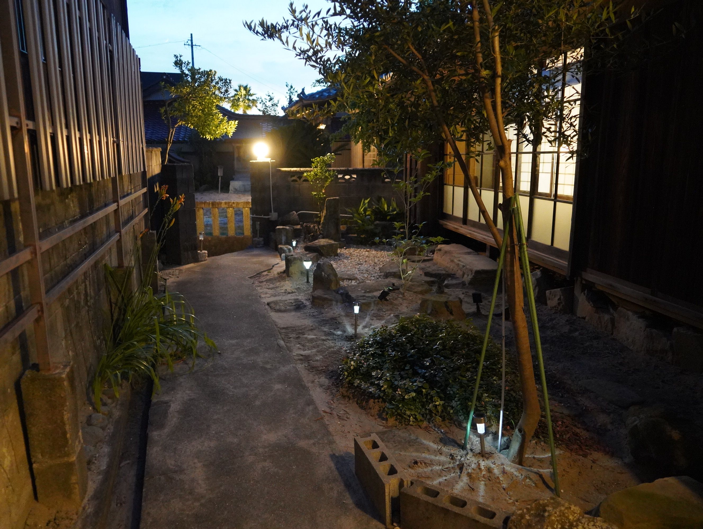 A narrow pathway flanked by stone structures and vegetation, leading to a traditional building with a tiled roof.