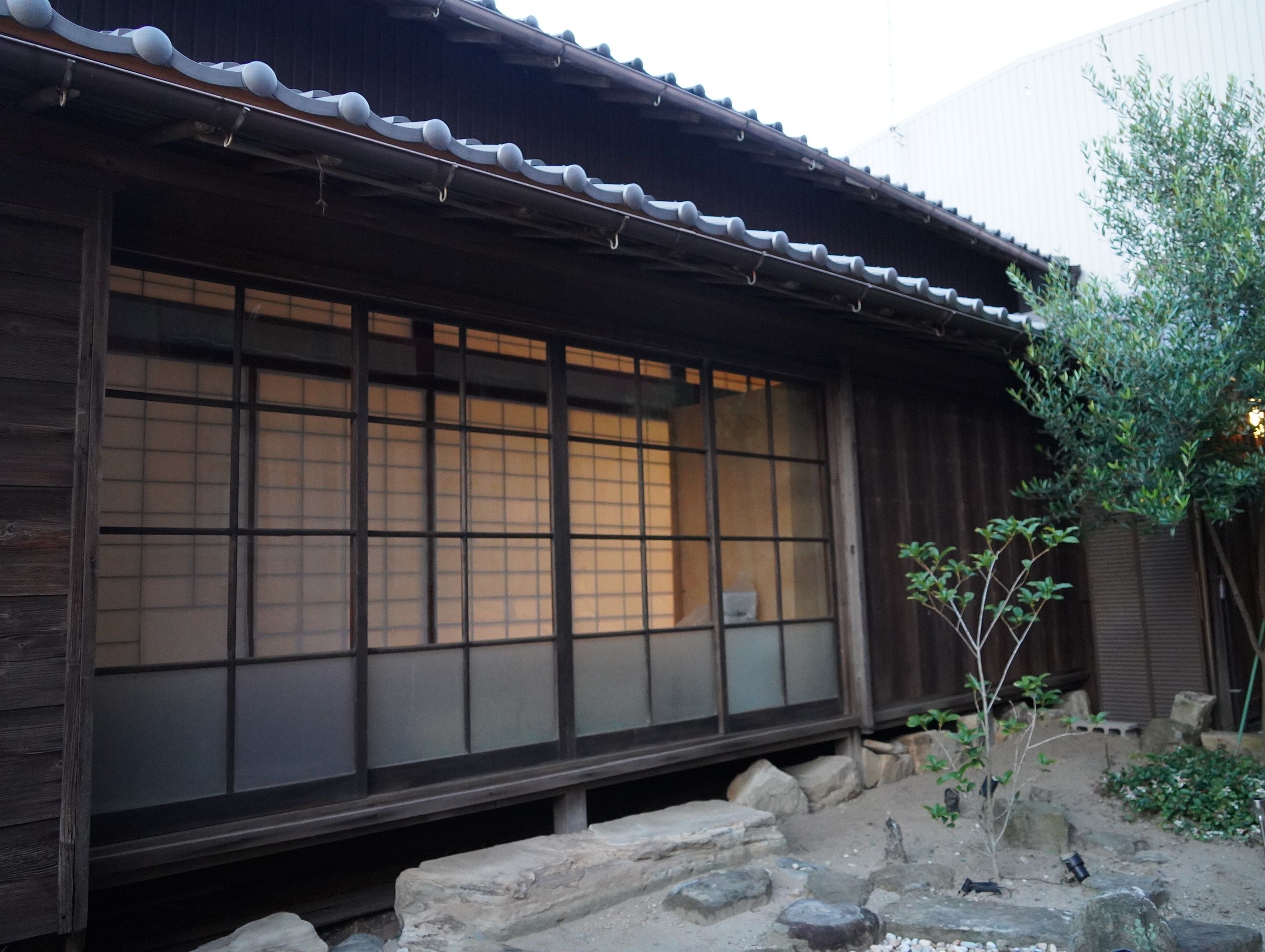 Un edificio tradicional japonés con puertas correderas, techo de tejas y un pequeño jardín con rocas y un árbol.