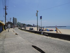 A coastal road with beachside buildings, a clear sky, and a beach with umbrellas and chairs. - New Listing (南知多町)