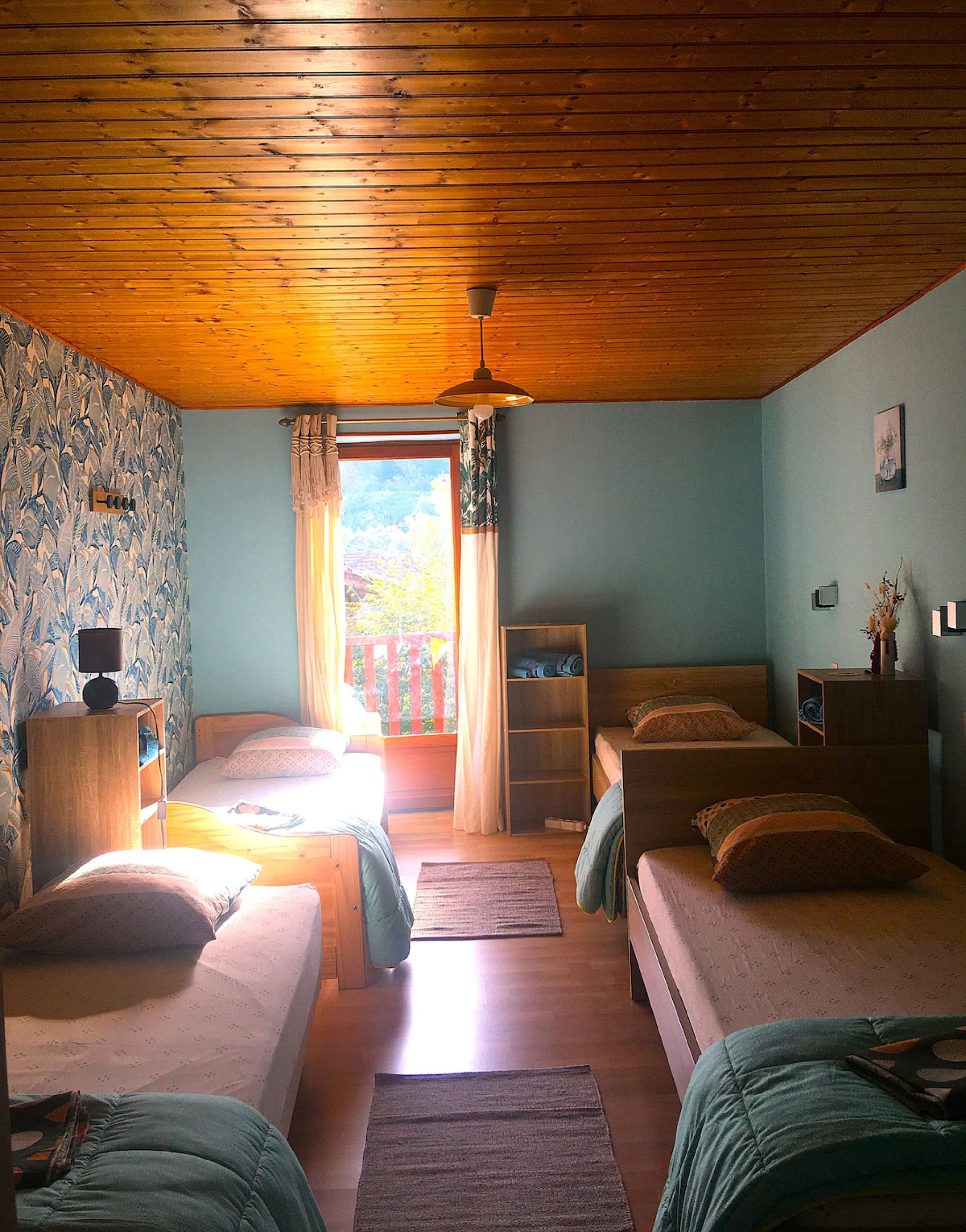 Une chambre avec deux lits, un plafond en bois, une fenêtre avec des rideaux, une petite table avec une lampe et un mur recouvert d’un papier peint à motifs.