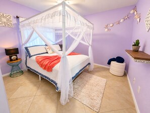 Ein Schlafzimmer mit einem Himmelbett, einem Nachttisch mit Lampe, einem kleinen Topfpflanzen und einem dekorativen Wandbild.