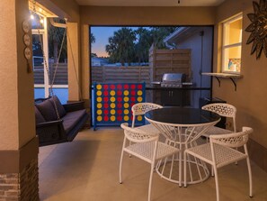Eine Terrasse mit einer Schaukel, einem runden Tisch mit Stühlen, einem Grill und einem bunten Gesellschaftsspiel.
