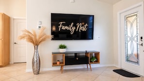 A modern living room with a flat-screen TV mounted on the wall, a black console table with plants, and a decorative vase with pampas grass. - Family Nest Pool&Gulf Coast Aventures (North Port)