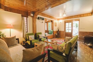 A cozy room with wooden walls and ceiling, a bed with a patterned bedspread, a green sofa with floral cushions, a glass coffee table, and a wooden side table with a lamp. - The Willow Way (Nainital)