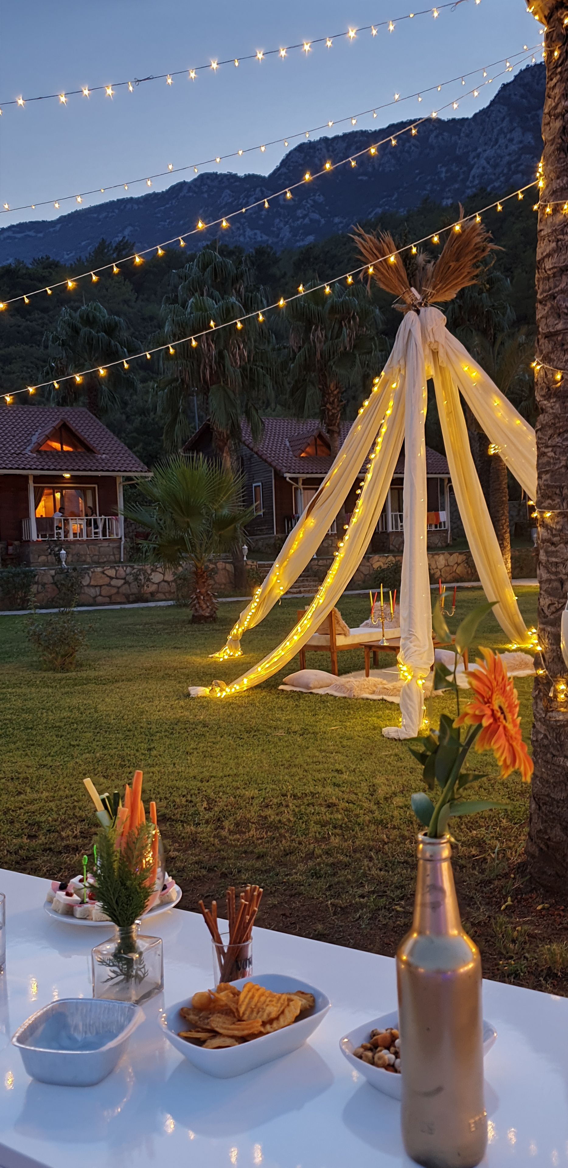 Un área de comedor al aire libre con luces colgantes, una mesa con aperitivos y una estructura parecida a una carpa.
