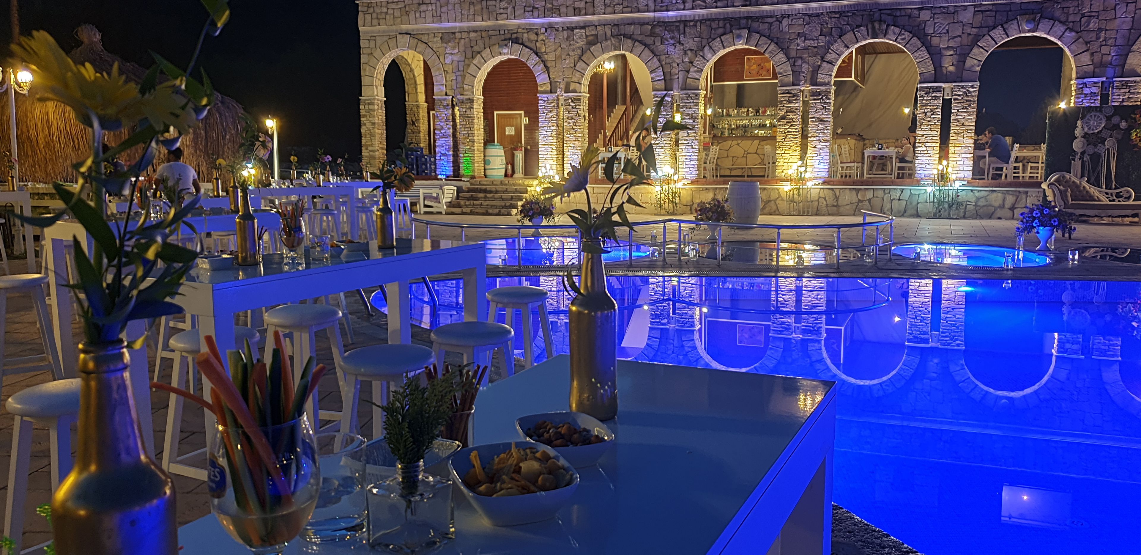 Zona de piscina exterior con arcos de piedra, asientos de bar y mesas con sombrillas.
