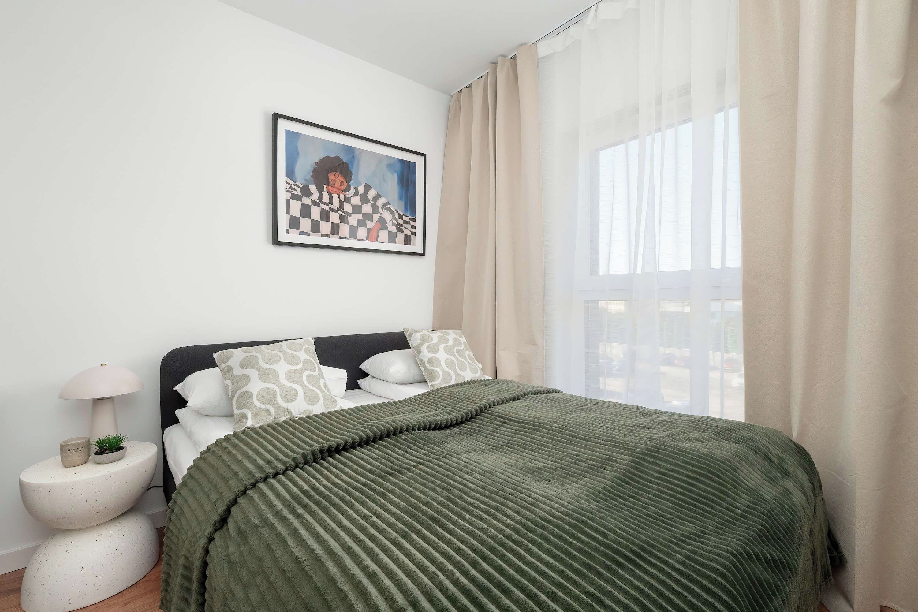 A bedroom with a bed, a bedside lamp, a potted plant, and a framed picture on the wall.