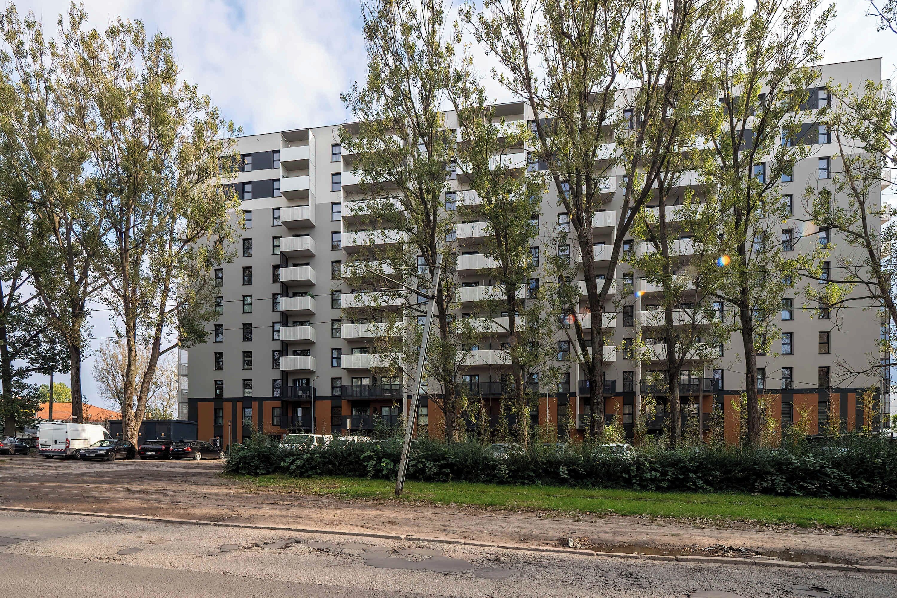 Een meerlaags woongebouw met balkons, omgeven door bomen en een parkeerplaats.