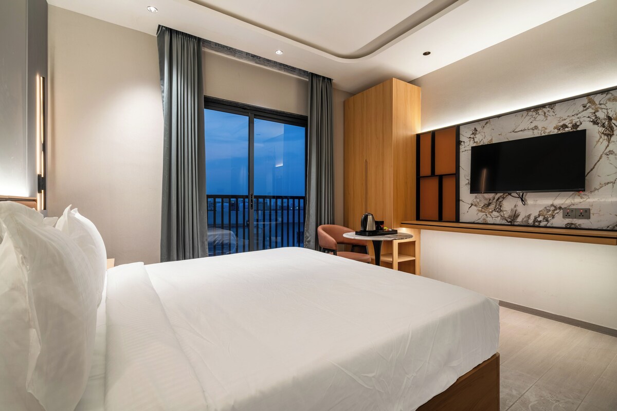 A hotel room with a bed, a small table with a kettle, a chair, a TV mounted on the wall, and a balcony with curtains.