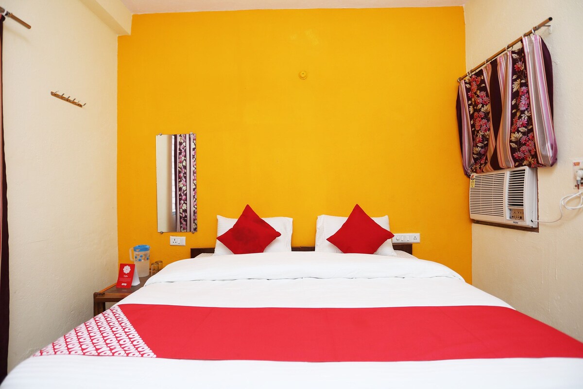 Une chambre d’hôtel avec un lit, deux oreillers rouges, un couvre-lit blanc, un mur jaune, un miroir, une fenêtre avec des rideaux, une table de chevet, une carafe d’eau et une prise électrique.