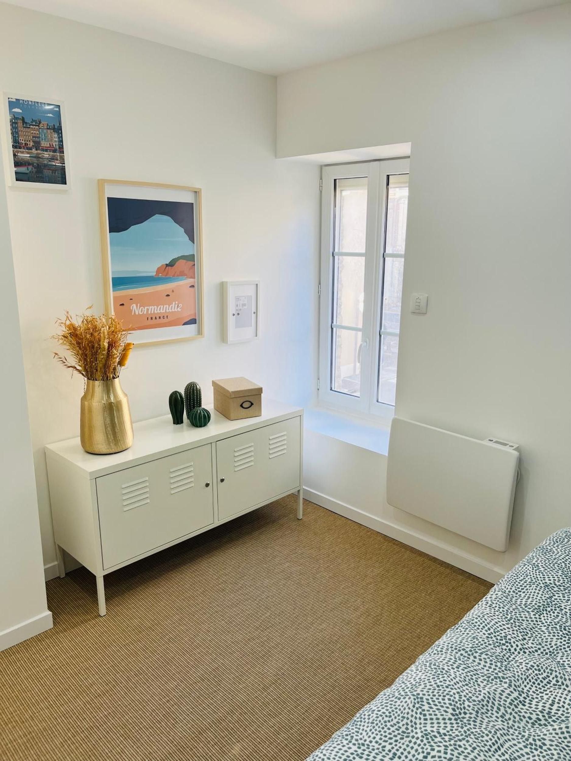 Ein Zimmer mit weißem Heizkörper, einem Fenster mit Jalousien, einem weißen Schrank mit Dekoration und einem gerahmten Bild an der Wand.