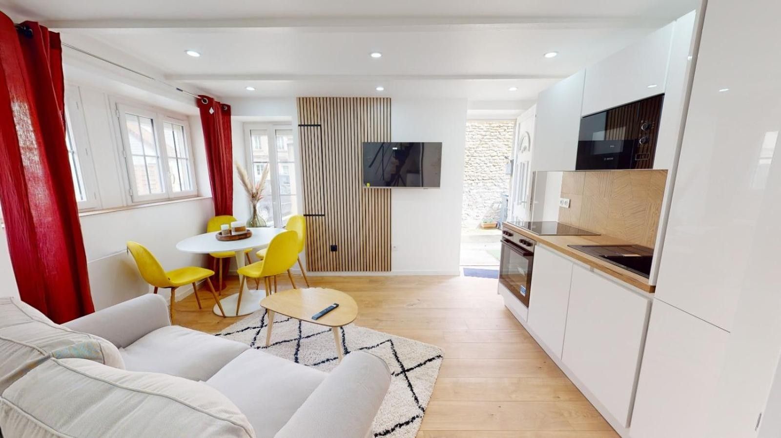 Een moderne woonkamer met een bank, een eettafel met gele stoelen, en een keuken met witte kasten en een aan de muur bevestigde televisie.
