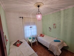 Huoneessa on kaksi sänkyä, pieni pöytä, jossa on lamppu, ja ikkuna, jossa on verhot.