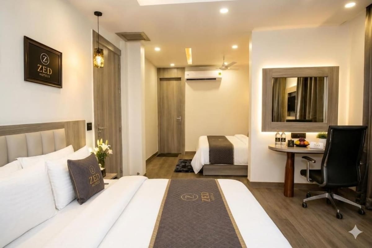 A hotel room with a bed, a desk, a chair, a mirror, and an air conditioning unit.