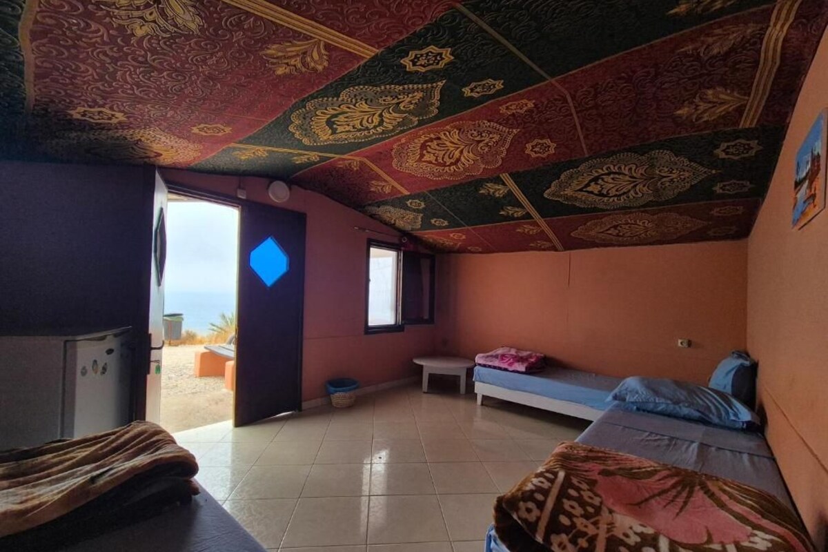 Ein Zimmer mit einem Bett, einer Bank, einem kleinen Tisch und Blick auf den Strand durch eine offene Tür.