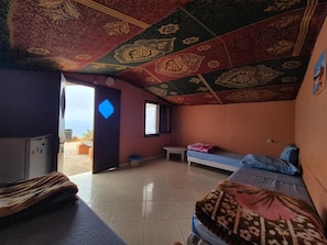 Ein Zimmer mit einem Bett, einer Bank, einem kleinen Tisch und Blick auf den Strand durch eine offene Tür.